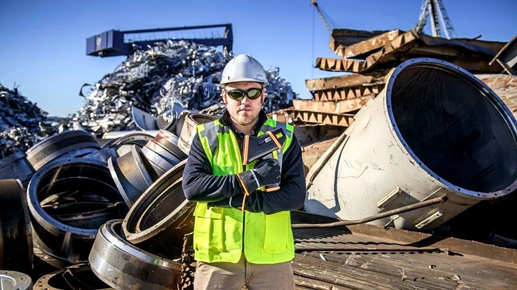 Man stands in front of a pile of metal scrap in a recycling yard.