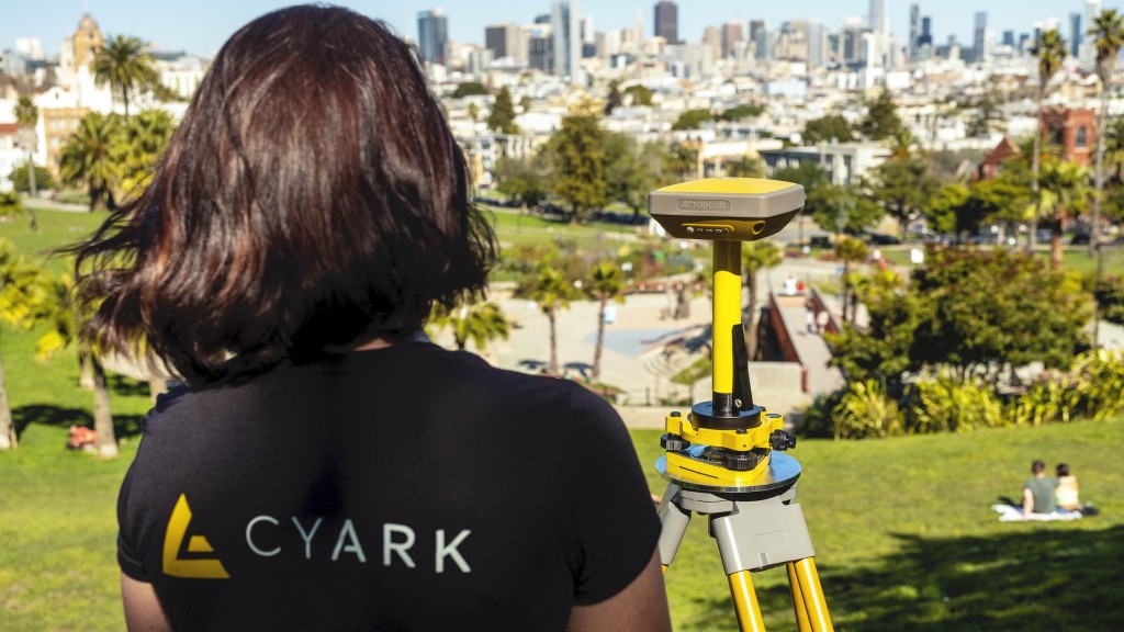 woman wearing CyArk t-shirt uses Topcon robotic total station on a hill looking over city