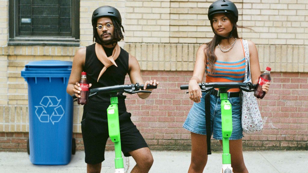 Two young electric scooter users hold Coca-Cola bottles on the street