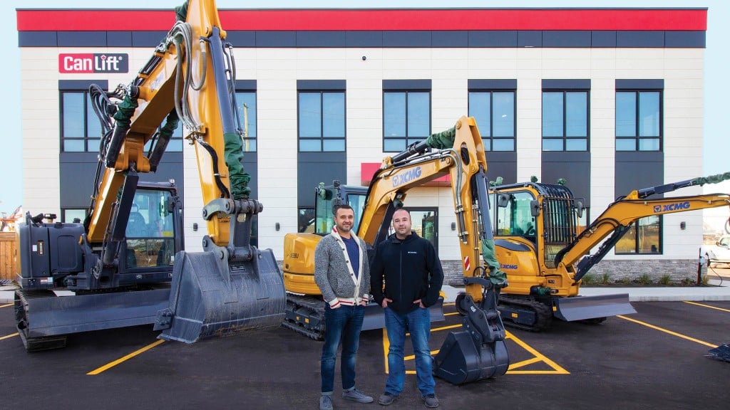 Two men stand in front of a line of XCMG machines and a dealership office