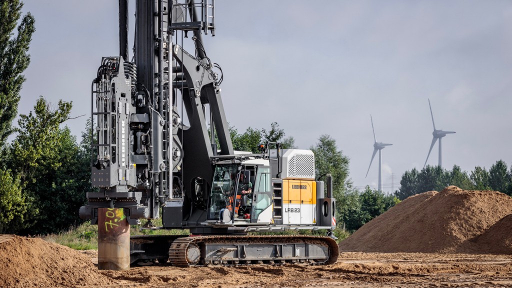 A piling rig drilling in a dirt field by a wind farm