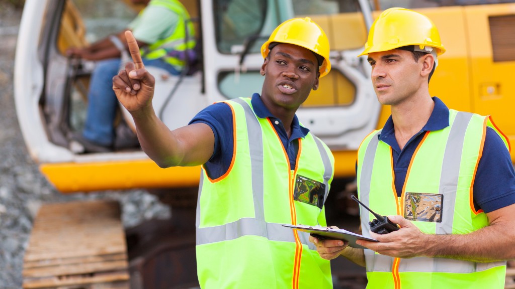 Two men stand in front of an excavator reviewing paperwork