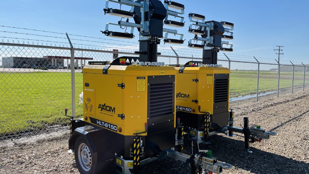 Two light towers are parked in a gravel parking lot near a fence