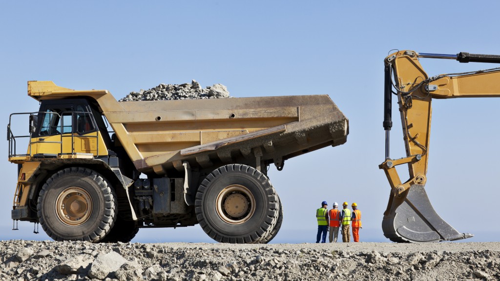 Workers stand on a mining job site near a truck and an excavator