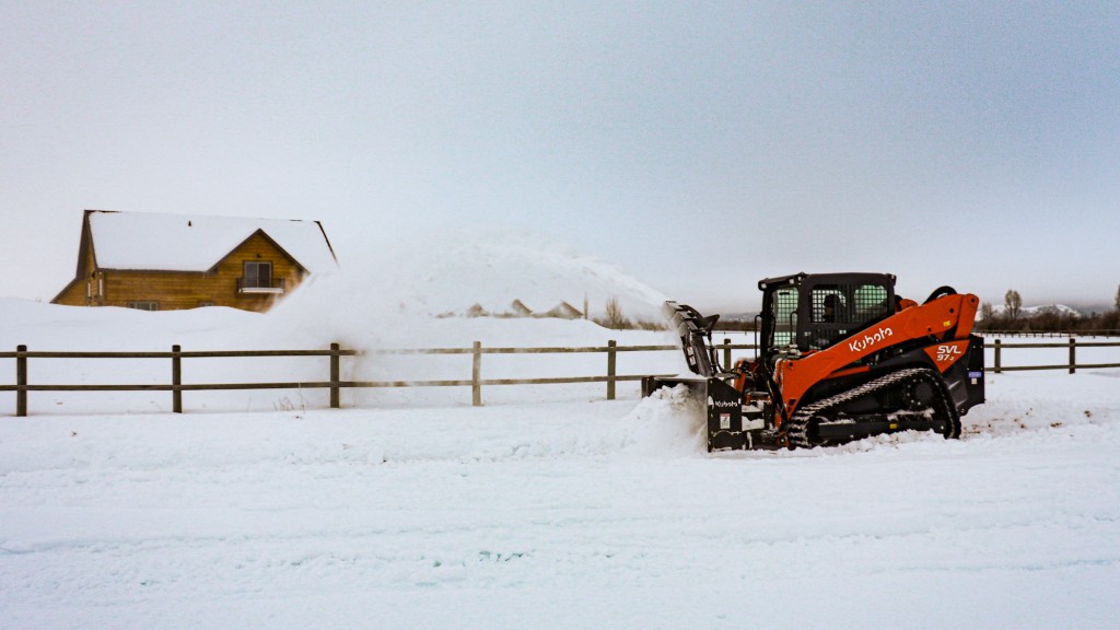 A compact track loader uses a snow blower on a snowy driveway