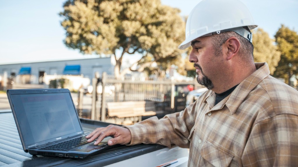 A man wearing a hard hat uses a laptop on a job site
