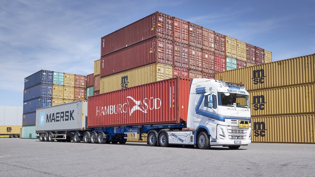 A truck with two trailers in front of piles of containers.