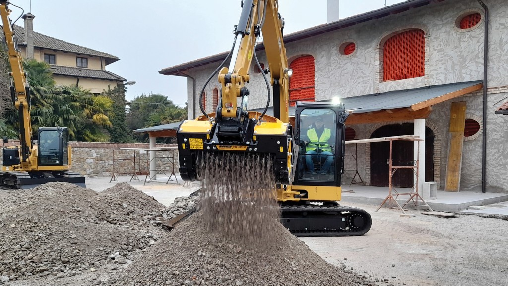 An excavator works with a crusher bucket attachment in front of a home.