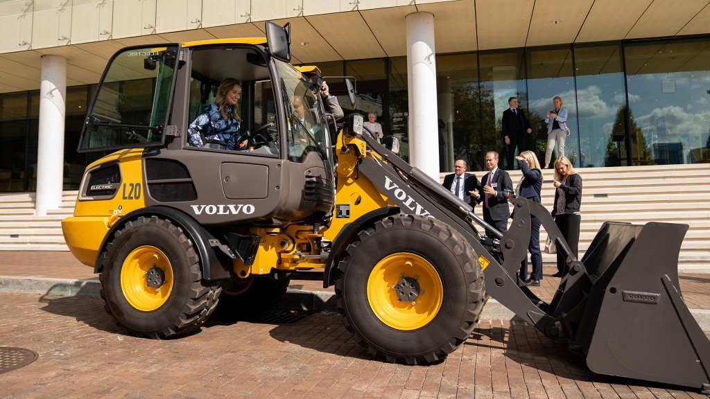 A woman sits in an electric wheel loader in front of a group of men in suits next to a concrete building.