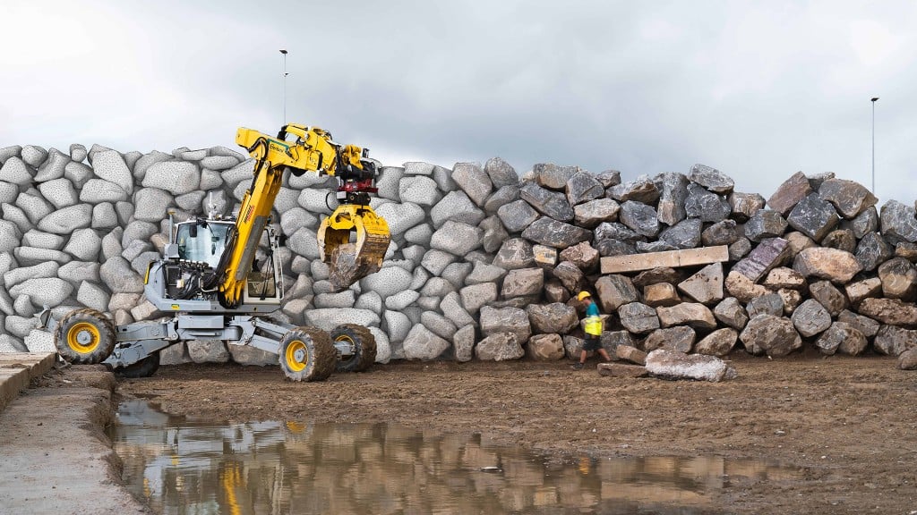 An excavator sits next to a large stone wall with a rock in its grapple.