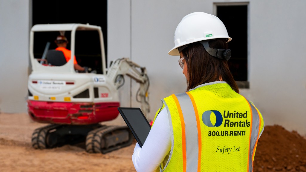 A woman in a hi-vis vest holding a tablet watches a mini excavator at work.