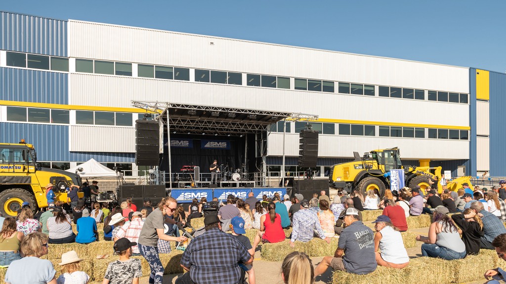 People sit watching a concert stage flanked by pieces of heavy equipment in front of a building.