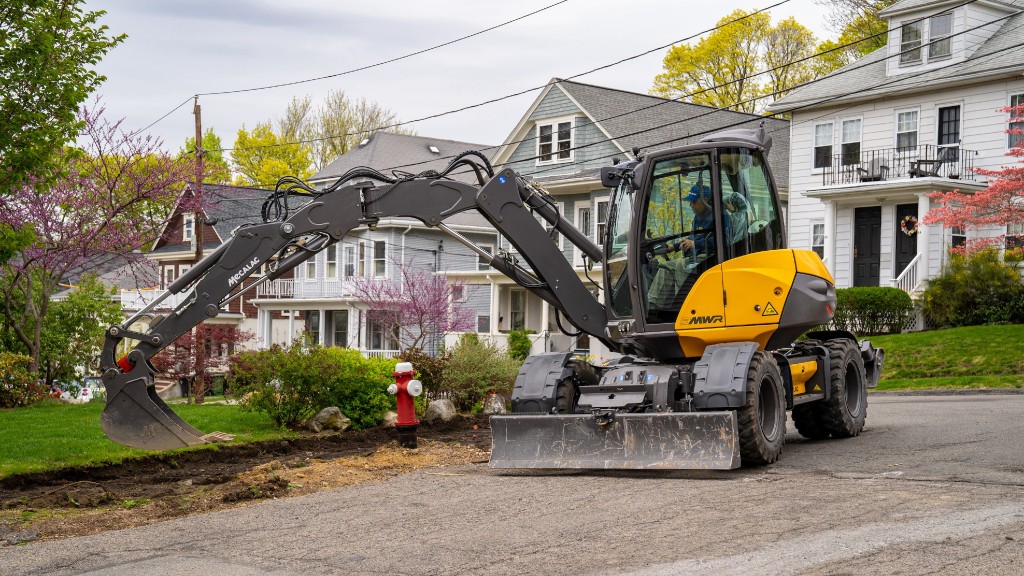 A compact wheeled excavator works near a residential driveway