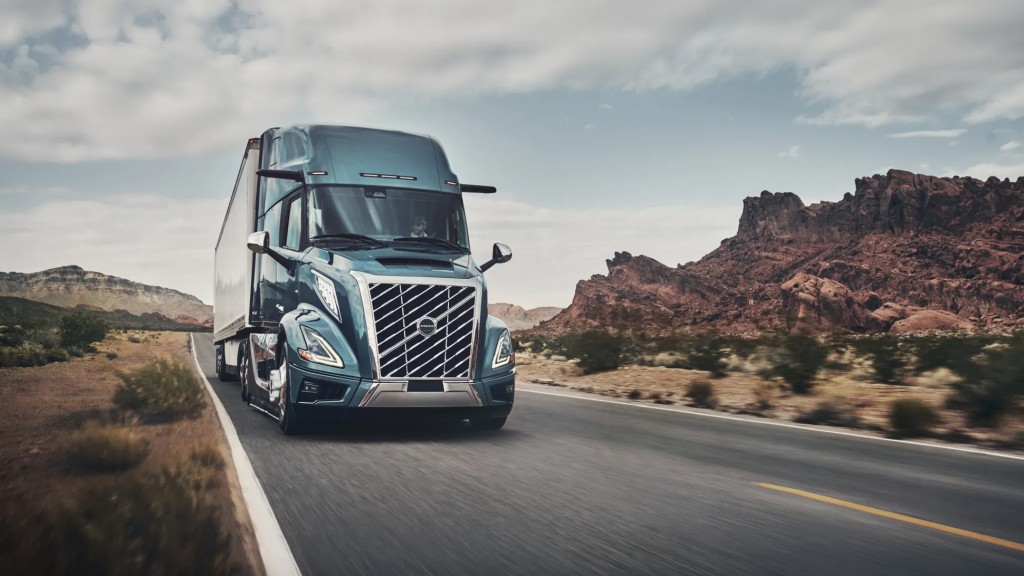 A blue semi truck towing a trailer on a road through an arid setting.