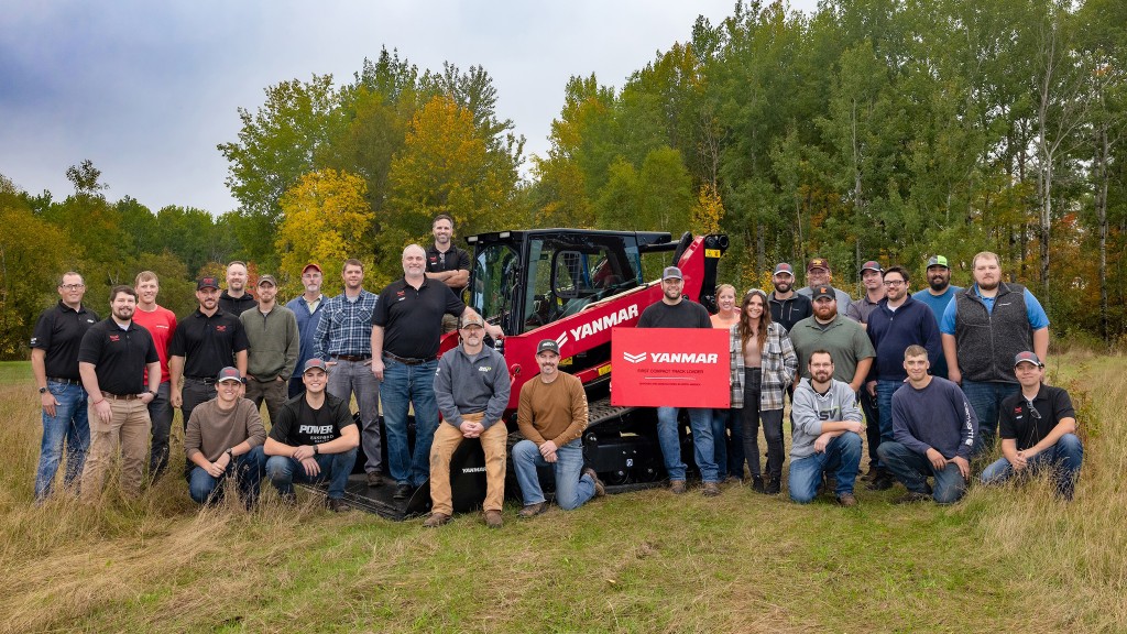 A group of people surrounding a red and black compact track loader on a grassy field.