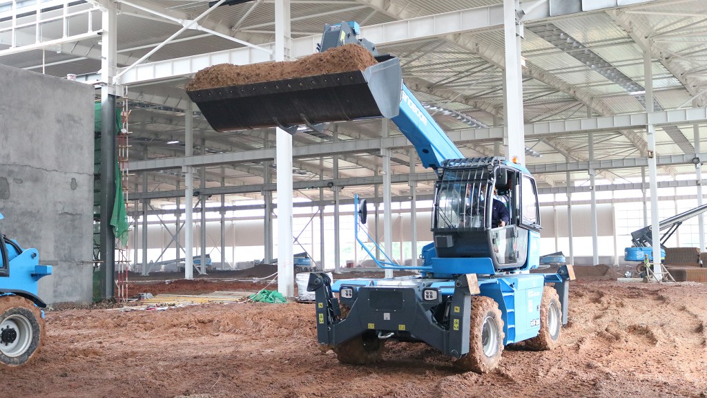 A blue telescopic handler with a bucket load of material inside a large structure.