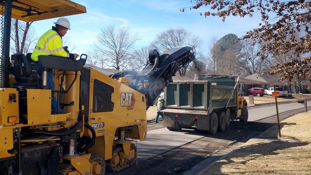 A cold planer places asphalt into a truck while working on a road.
