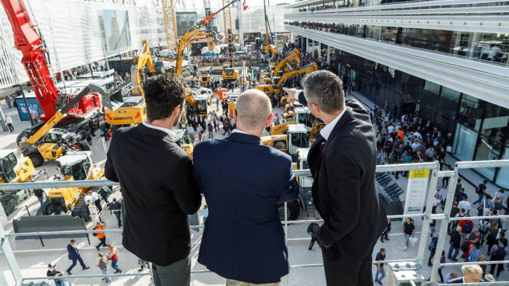 Three people on a raised walkway look out over a display of Liebherr equipment in a large trade show booth.