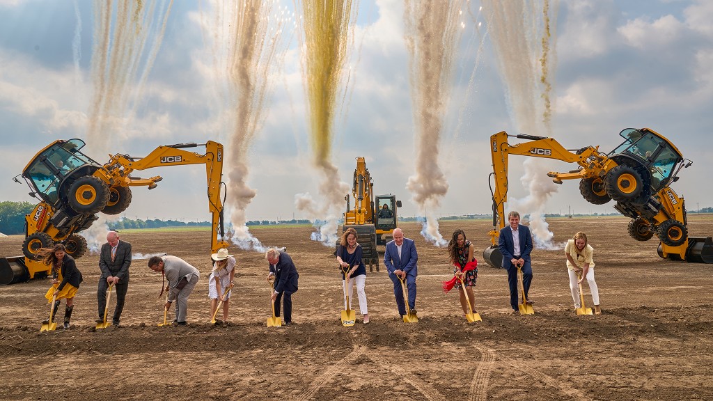 A line of people use ceremonial shovels to break ground, in front of two backhoes and an excavator and a smoke show.