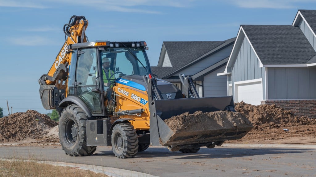 An electric backhoe loader carries a bucket full of dirt down a road