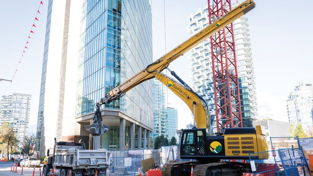 A telestick excavator dumps dirt from a clamshell bucket into a dump truck