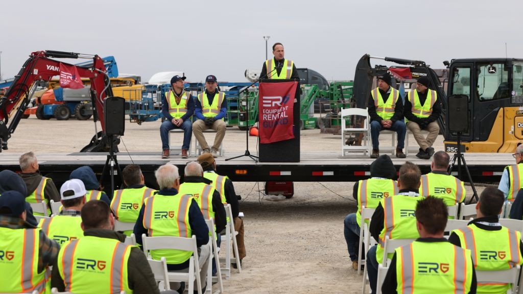 A man stands at a podium while several men sit in the audience, all are wearing high visibility vests A man stands at a podium while several men sit in the audience, all are wearing high visibility vests