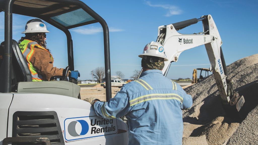 A woman operating a compact backhoe loader with a man next to her A woman operating a compact backhoe loader with a man next to her