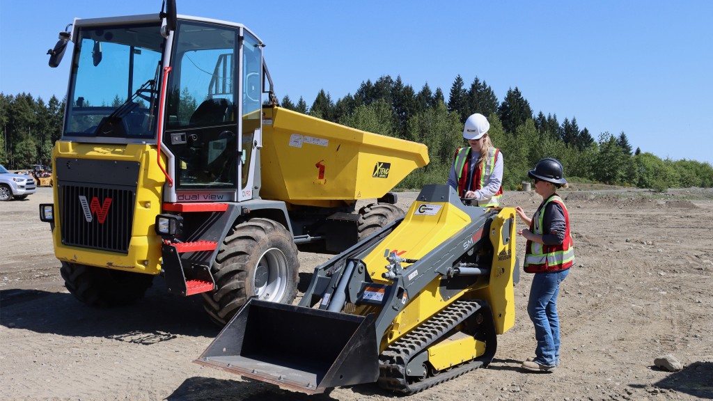 A female student operates a compact track loader under the guidance of a female instructor on a work site. There is a a mini dumper behind them. A female student operates a compact track loader under the guidance of a female instructor on a work site. There is a a mini dumper behind them.