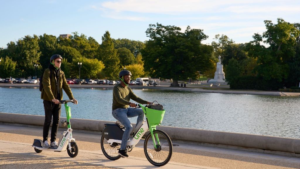 Two people driving an e-scooter and e-bike next to a small lake Two people driving an e-scooter and e-bike next to a small lake
