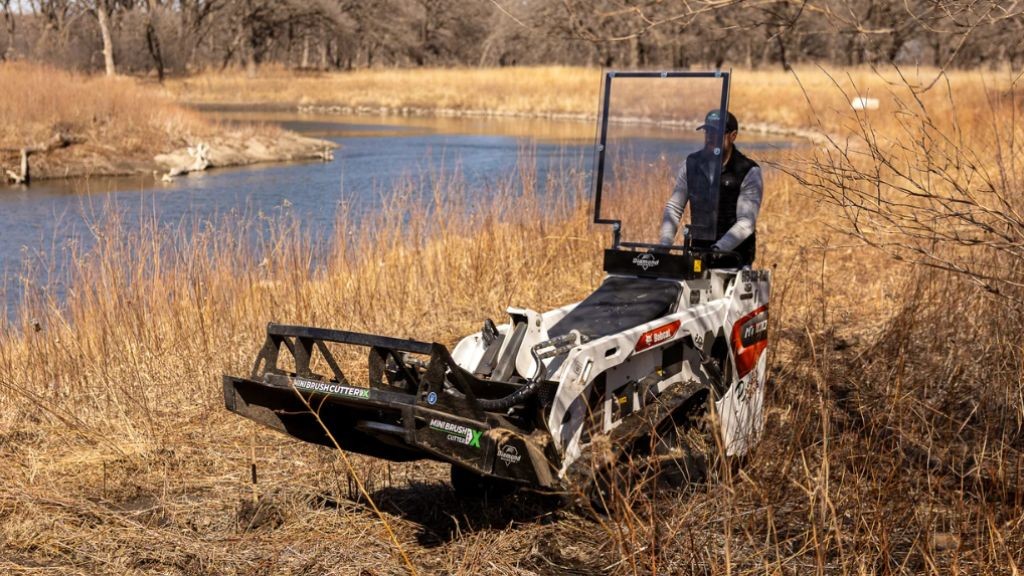 A man on a riding mower moving through dry grass next to a small river