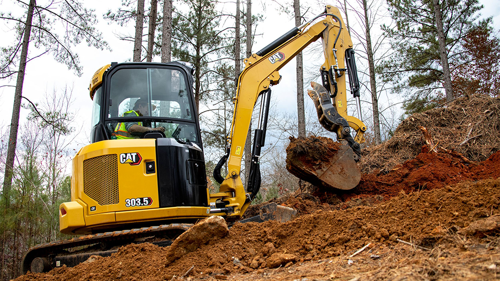 A mini excavator on a small hill surrounded by trees A mini excavator on a small hill surrounded by trees