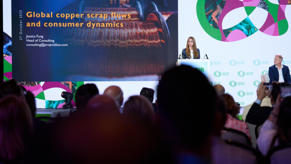 A woman standing at a podium on a stage with a large presentation behind her in front of a seated audience