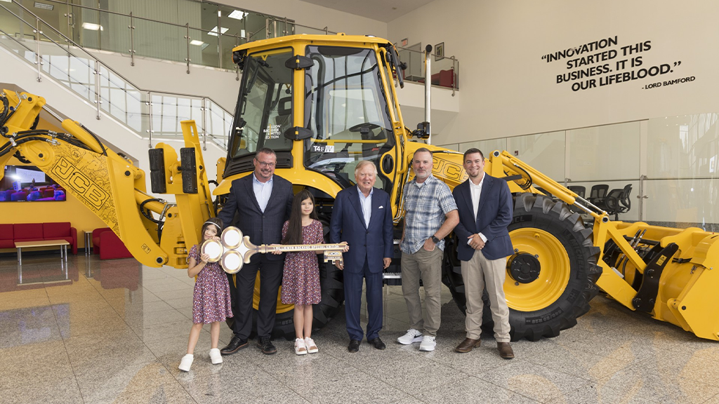 A group of people posing for a photo in front of a large yellow backhoe loader A group of people posing for a photo in front of a large yellow backhoe loader