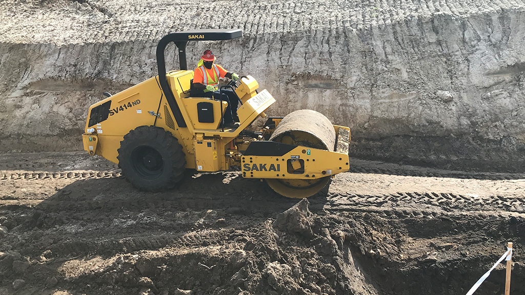 A man operates a single drum soil compactor on an outdoor site