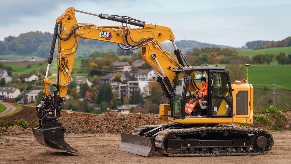 A yellow excavator sits on dirt with a town behind it in the hills