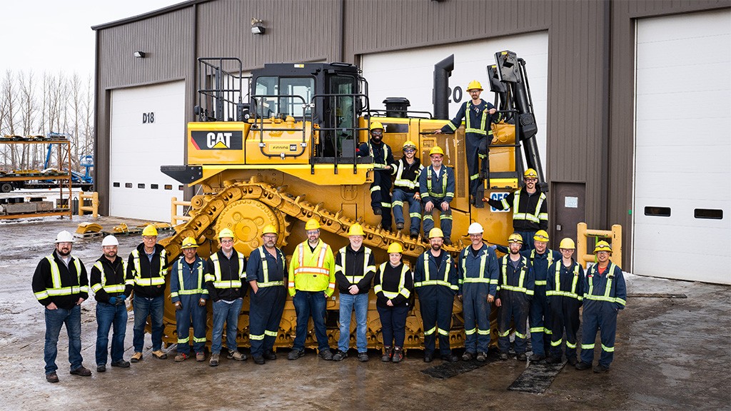 A team of heavy equipment mechanics wearing coveralls take a group photo in front of a large dozer