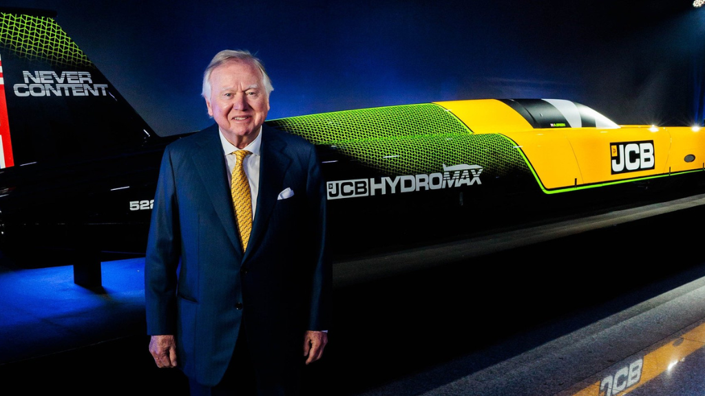 A man wearing a suit standing in front of a race car with the JCB Hydromax logo and a yellow and green paint scheme