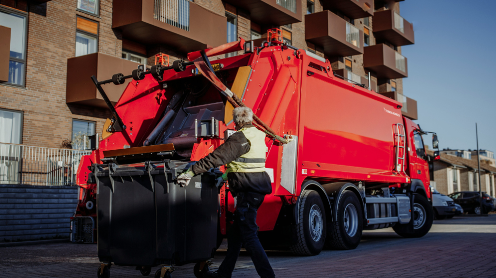 A person rolls a residential waste bin closer to the back of a red waste hauling vehicle A person rolls a residential waste bin closer to the back of a red waste hauling vehicle