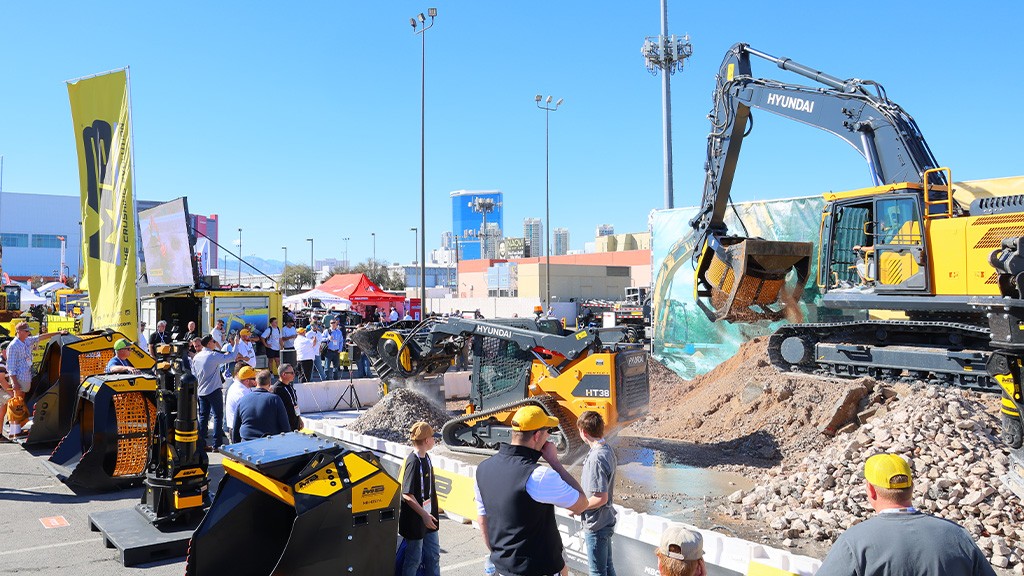 An excavator manoeuvres through an outdoor site surrounded by people and attachments An excavator manoeuvres through an outdoor site surrounded by people and attachments