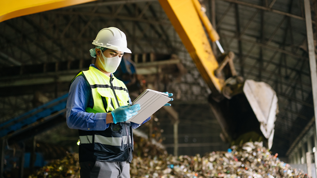 A man wears PPE and holds a clipboard in front of a large pile of solid waste and an excavator bucket A man wears PPE and holds a clipboard in front of a large pile of solid waste and an excavator bucket