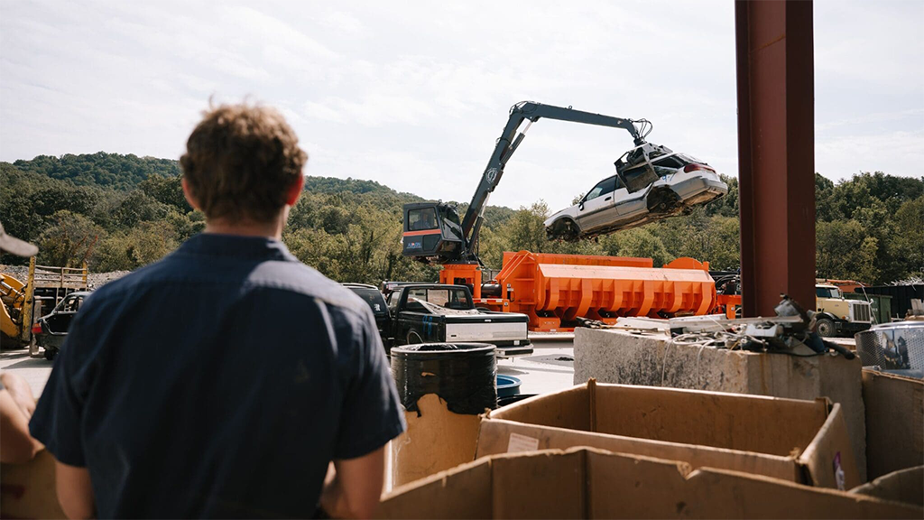 A man stands in front of an end-of-life car being lifted by a material handler A man stands in front of an end-of-life car being lifted by a material handler