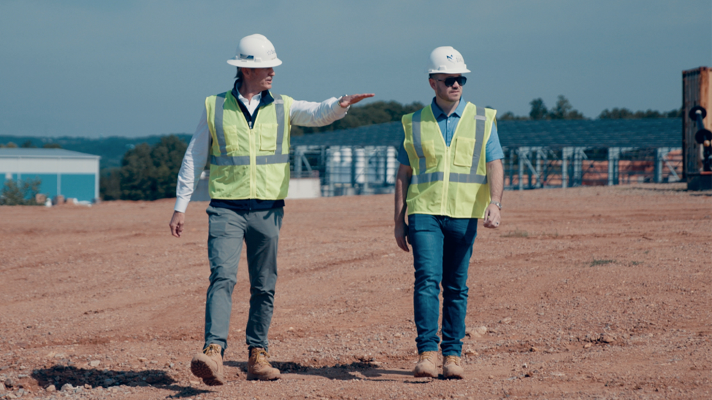Two men wear PPE while walking across a site Two men wear PPE while walking across a site