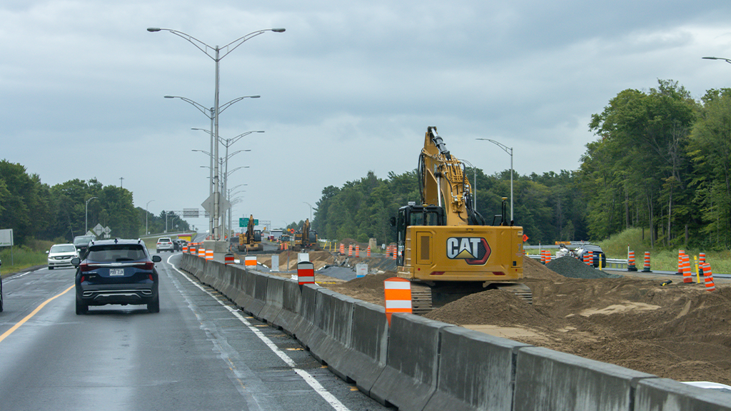 A Caterpillar excavator works on the side of a highway while cars drive by A Caterpillar excavator works on the side of a highway while cars drive by