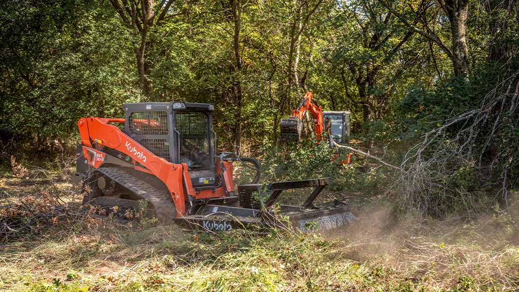Two compact machines operate attachments in a forested area Two compact machines operate attachments in a forested area